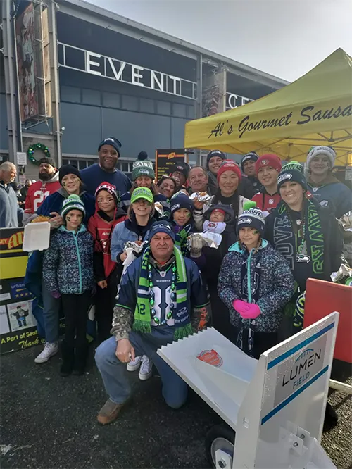 A group of smiling people in Seahawks gear pose outside Lumen Field near a food stand.