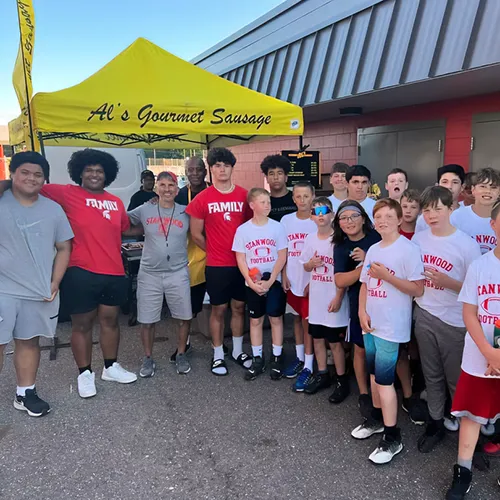 A group of teens and adults pose together outside under a yellow "Al's Gourmet Sausage" tent.