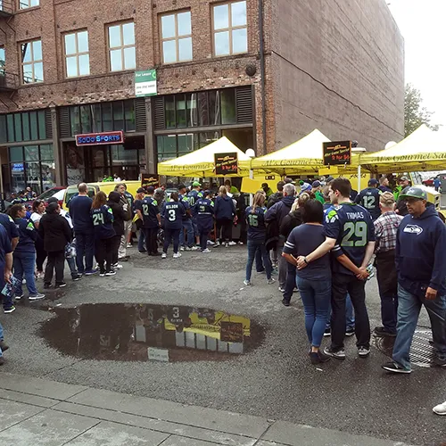 A crowd in sports jerseys gathers outside food stalls by a brick building on a cloudy day.