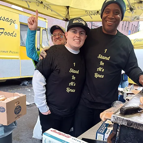 Two smiling men wear matching "I Stand In Al's Lines" shirts at an outdoor food stand, with a yellow truck behind.