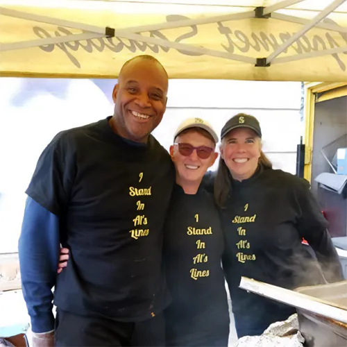 Three smiling people in matching “I Stand In Al’s Lines” shirts stand together under an outdoor canopy.