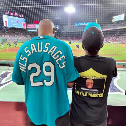 Two people at a baseball game, one wearing an “Al’s Sausages 29” jersey, the other a custom sausage t-shirt.