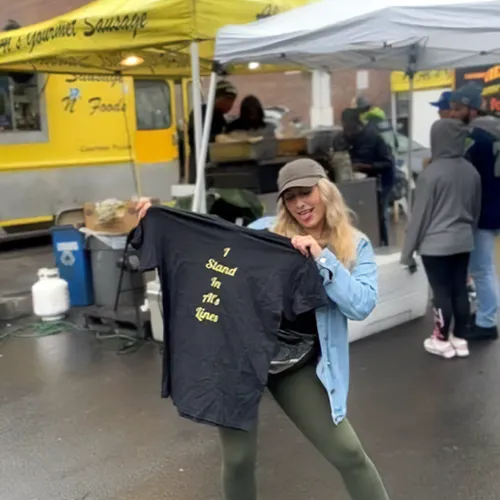 Woman holding a black T-shirt that says "I Stand In The Lines" at an outdoor food market.