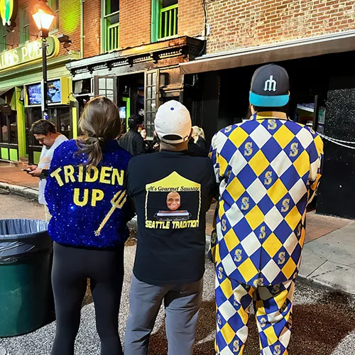 Three people in colorful sports-themed outfits stand with their backs to the camera on a city sidewalk at night.