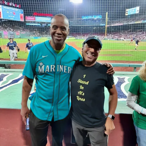 Two men smiling at a baseball stadium; one wears a Mariners jersey, the other a black T-shirt with yellow text.