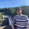 A person in a striped shirt poses in front of a waterfall and forest under a clear blue sky.