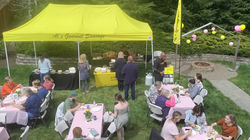 People eat at outdoor tables near a yellow tent labeled “Al’s Gourmet Sausage” at a garden party.