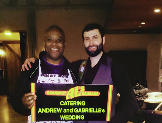 Two smiling men hold a "Gourmet Al's Sausage" sign for Andrew and Gabrielle's wedding.