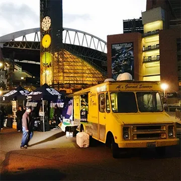 A yellow food truck parked near a stadium at dusk, with people and tents nearby.