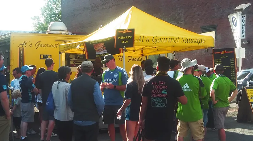 A crowd stands in line at Al’s Gourmet Sausage yellow food truck on a sunny day.