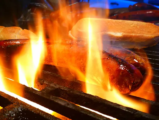 Sausage grilling over open flames with buns in the background.