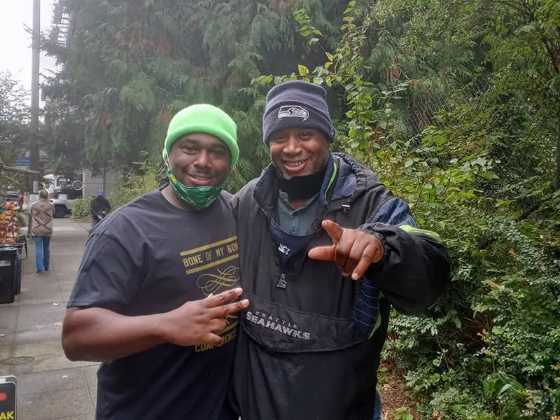 Two men smiling and posing together outdoors in winter clothes, surrounded by greenery.