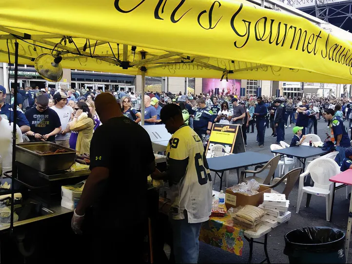Two people serve food under a yellow tent at a crowded outdoor event with sports fans in jerseys.