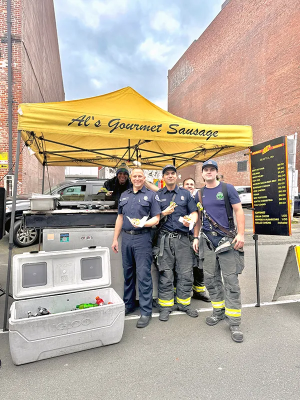 Four firefighters stand in front of Al’s Gourmet Sausage yellow tent, holding food and smiling.