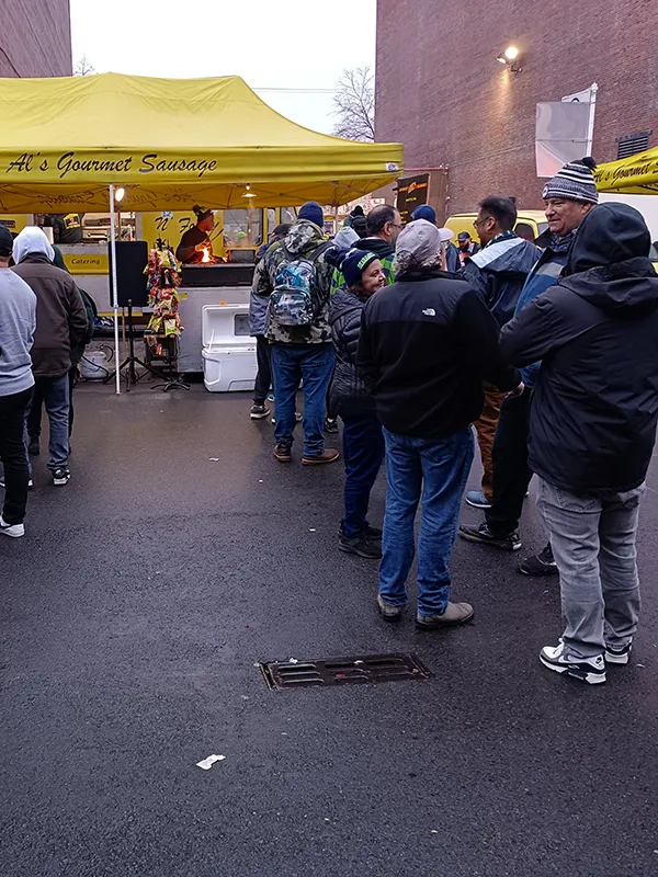 People stand in line at a yellow Al’s Gourmet Sausage food tent between two brick buildings on a cloudy day.