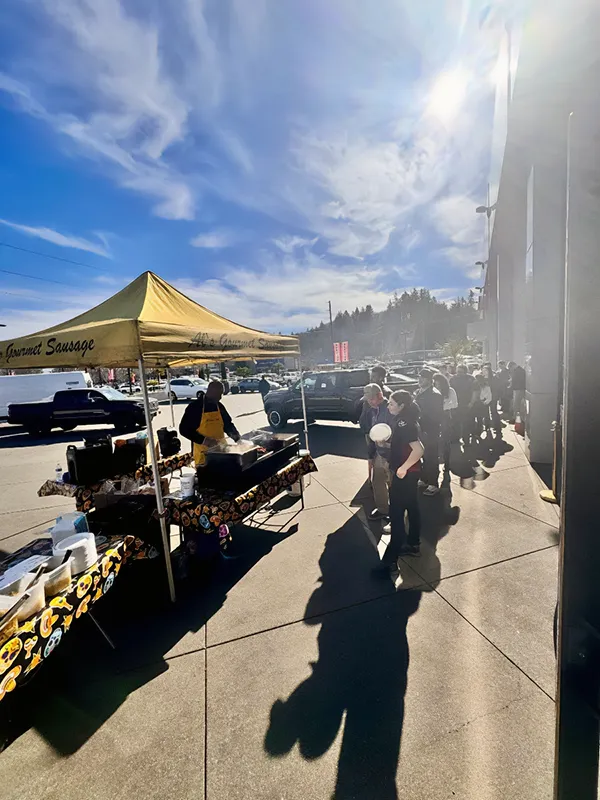 People line up at a yellow food tent labeled "Gourmet Sausage" on a sunny day in a parking lot.