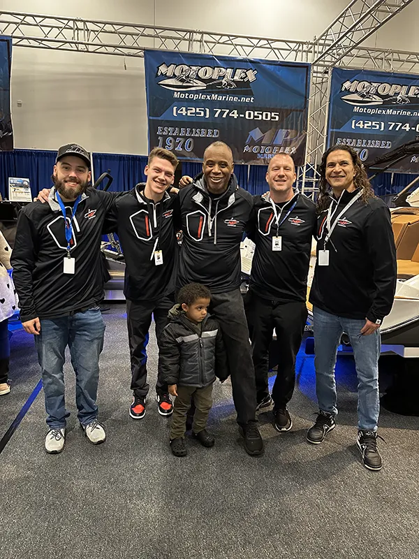 Five men in matching jackets pose and smile at a boat show, with a young child standing in front of them.