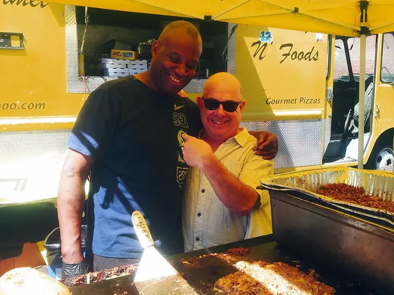 Two men smiling together at an outdoor food stand, with grills and trays of food in front of them.