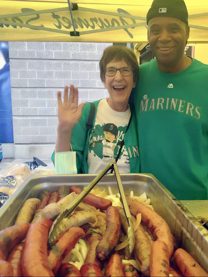 Two people smile at a food stand with grilled sausages in front, under a Mariners tent. One person is waving.