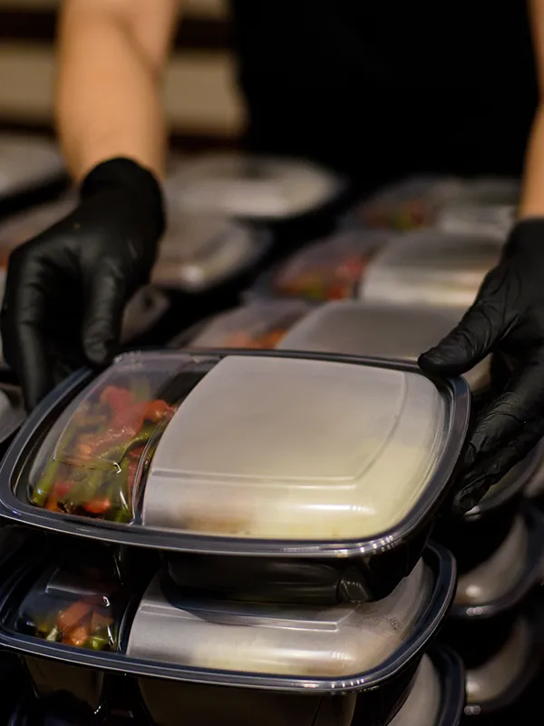 Gloved hands arrange stacks of closed takeaway meal containers with food inside.