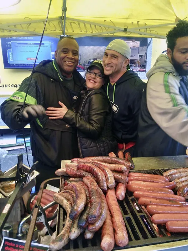 Three people smile and pose together behind a grill filled with sausages and hot dogs at an outdoor event.