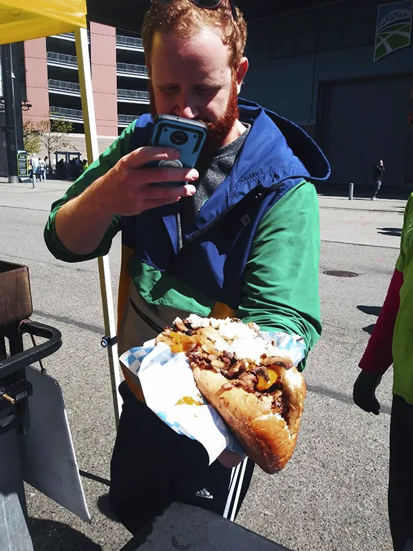 A man takes a photo of a large, overstuffed sandwich with his phone at an outdoor event.
