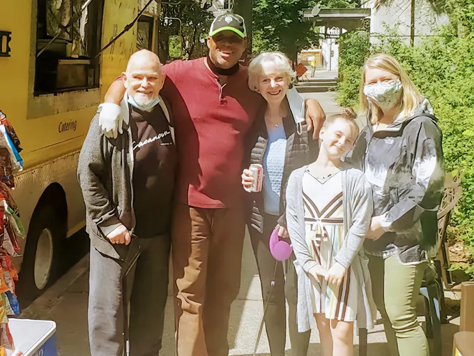 Five people smile together on a sunny sidewalk near a yellow food truck, with trees in the background.