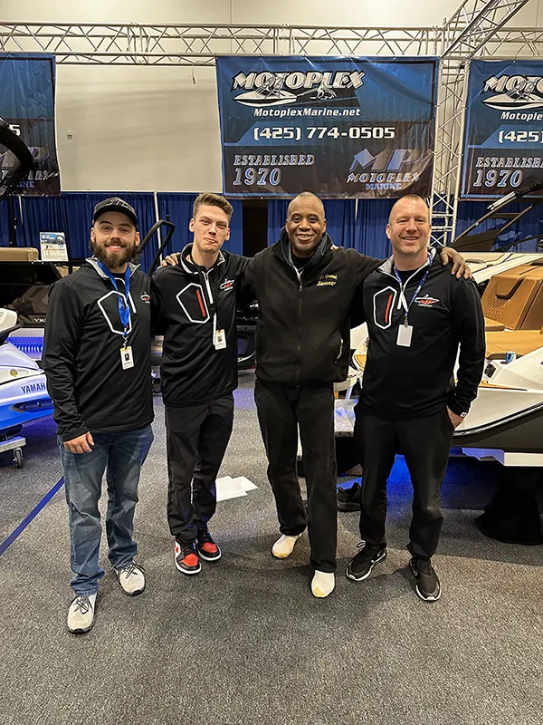 Four men stand together smiling at a Motoplex Marine event with boats and banners in the background.