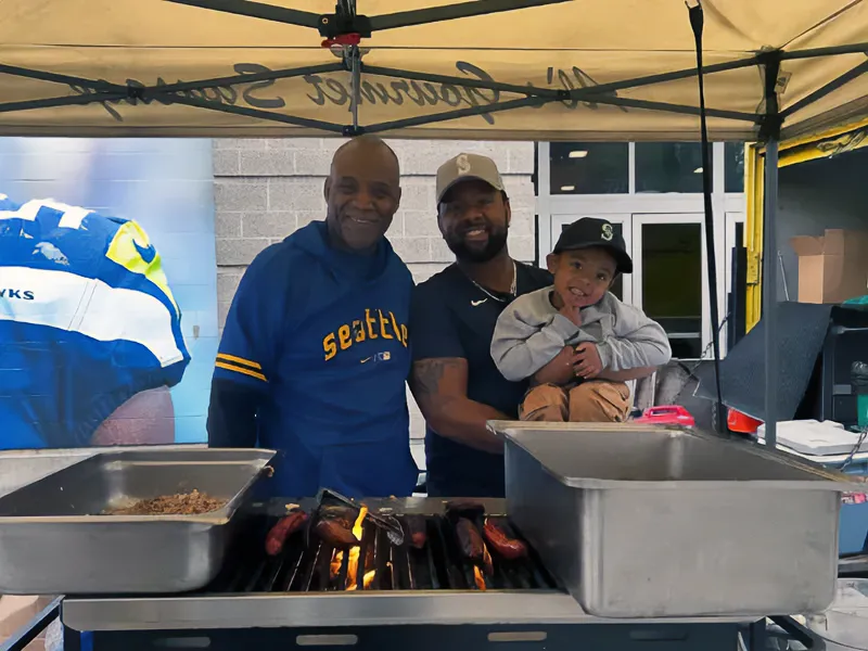 Two men and a child smiling at a grill stand under a Seattle-themed tent, with food cooking on the grill.