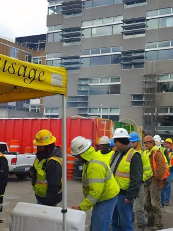 Construction workers in safety gear line up by a yellow tent at an urban construction site.