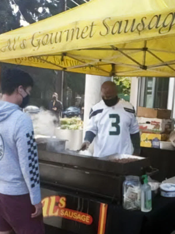 A vendor grills food at a sausage stand while a customer waits under a yellow tent at an outdoor event.