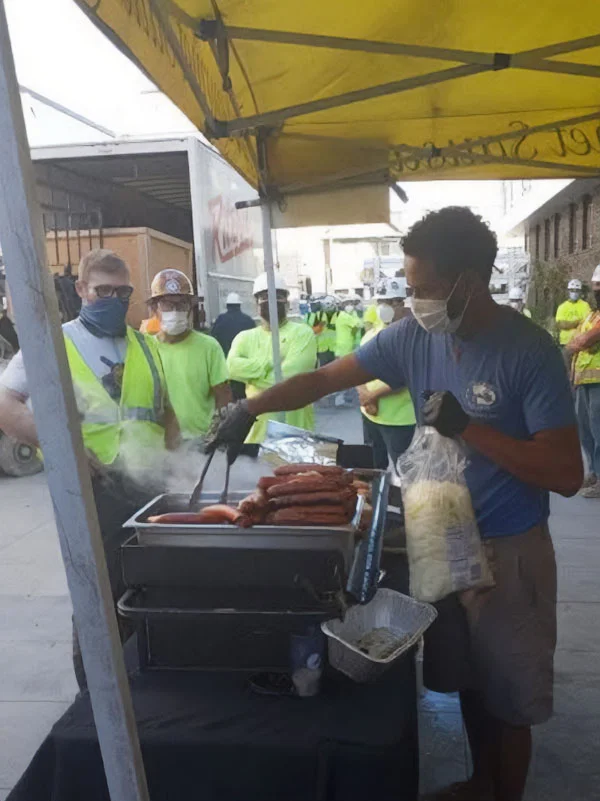 A man serves hotdogs under a yellow tent while workers in safety vests wait in line outside.