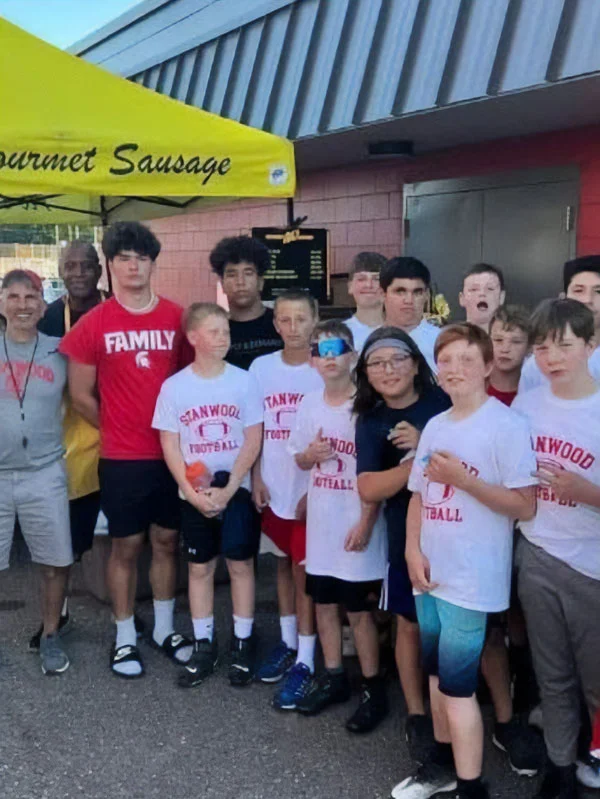 Group of boys in Stanwood Football shirts pose with adults under a yellow tent labeled "Gourmet Sausage.