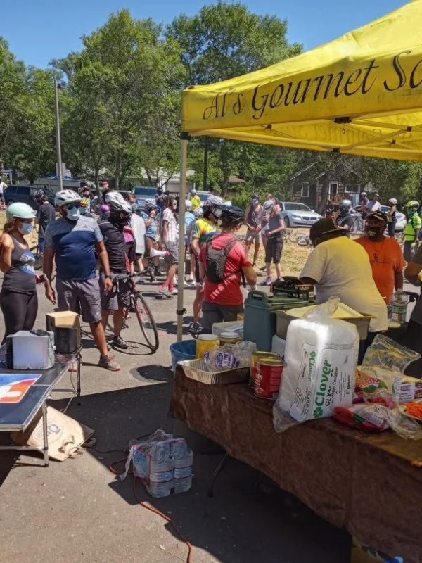People gather under a yellow tent at an outdoor event with food and supplies on tables, many wearing helmets.