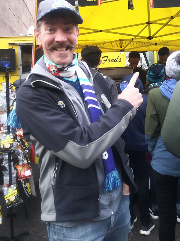 Smiling man in a cap and scarf points at a food stall, with people gathered around under a yellow tent.