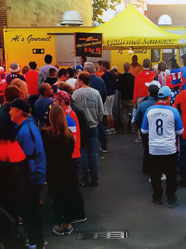 People line up at a yellow gourmet sausage food truck at an outdoor event, some wearing sports jerseys.