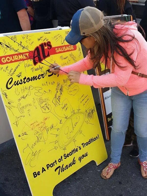 Woman in pink hoodie signs a large yellow board covered with signatures at a food stand.