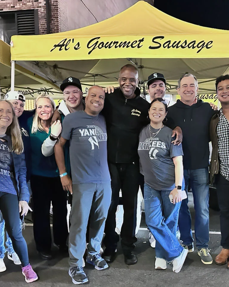 A group of people smiling together in front of a yellow "Al's Gourmet Sausage" tent at night.