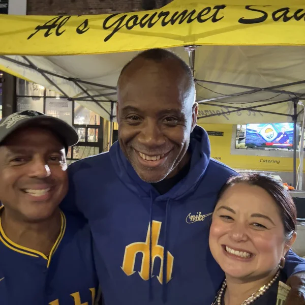 Three people smiling together in front of a yellow tent with "Al's Gourmet" written on it.