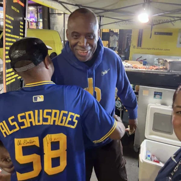 Two men happily greet each other near a food cart; one wears a “Sausages 58” jersey.